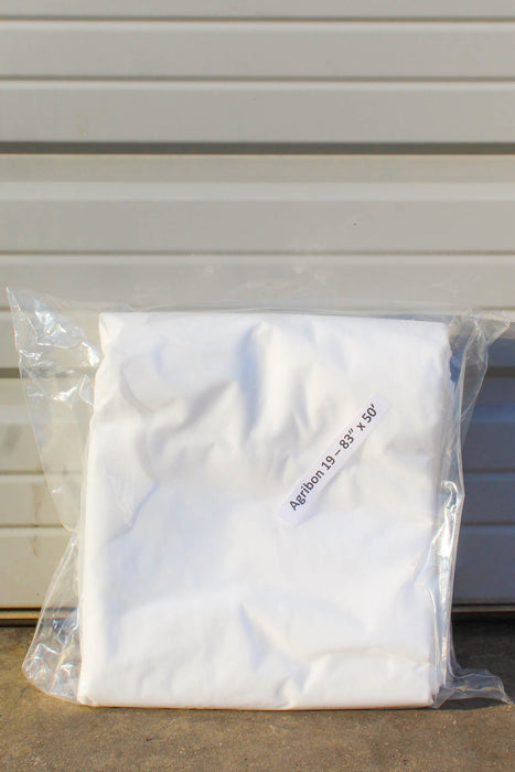 A folded white fabric item sealed in a clear plastic bag, with small printed text reading 'Agribon 19 83" x 50' on the bag, photographed against a light gray corrugated wall.