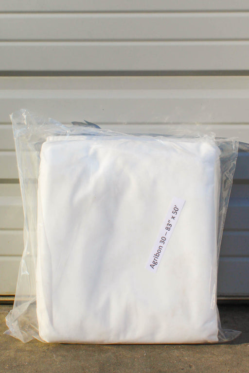 A folded white fabric item, sealed inside a clear plastic bag with a printed size label reading 'Agribon 30  83" x 50'. The bag is placed on a concrete surface against a gray corrugated metal wall.