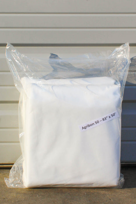A folded white fabric sealed in a clear plastic bag, with a small label reading 'Agribon 50  83" x 50' on a light siding background.