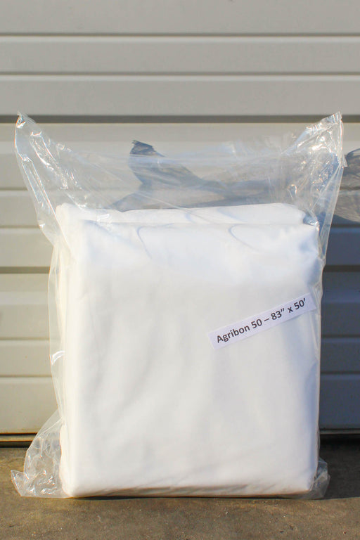 A folded white fabric sealed in a clear plastic bag, with a small label reading 'Agribon 50  83" x 50' on a light siding background.