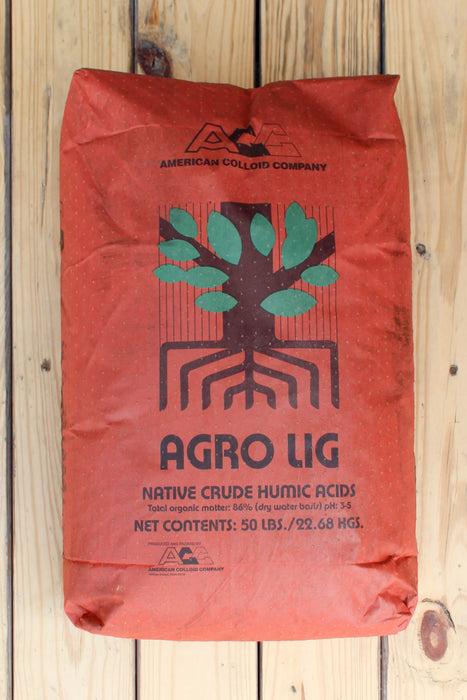 A 50 lb bag of American Colloid Company Agro Lig Native Crude Humic Acids, in a textured orange-red bag with a green tree and brown roots logo, placed on a light wood plank background.