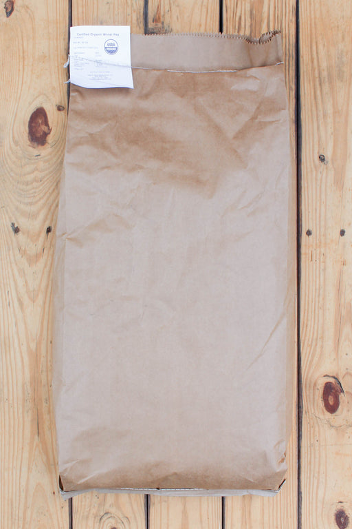 A brown kraft paper bag of Austrian winter peas with a white label, laid flat on a light-colored wooden plank surface.
