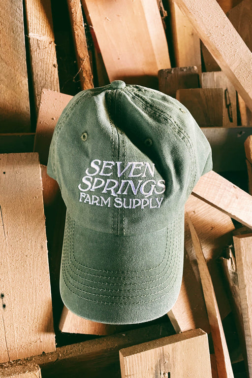 An olive green baseball cap with 'SEVEN SPRINGS FARM SUPPLY' embroidered in white on the front, resting on a stack of wooden pallets.