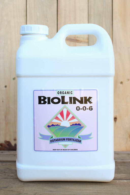 A white plastic jug of Organic BioLink 0-0-6 Potassium Fertilizer, featuring a colorful label with a sun and wave graphic, set against a rustic wooden background.