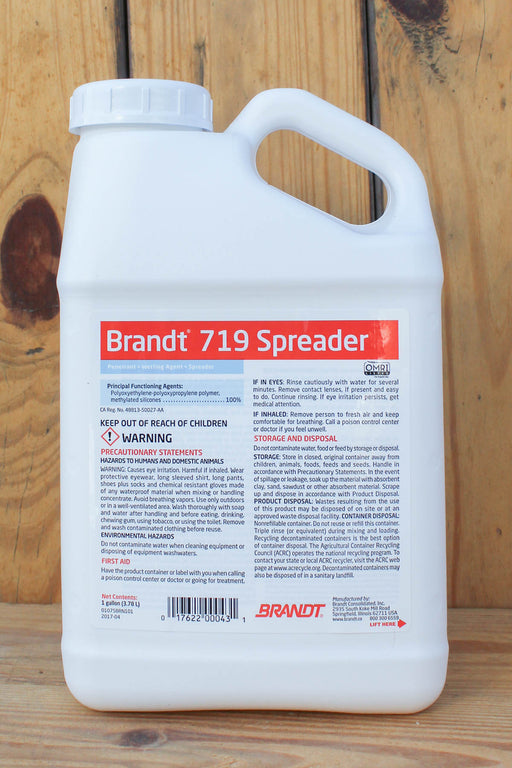 A white plastic jug of Brandt 719 Spreader with a detailed product label, photographed standing upright on a light-colored wooden surface.