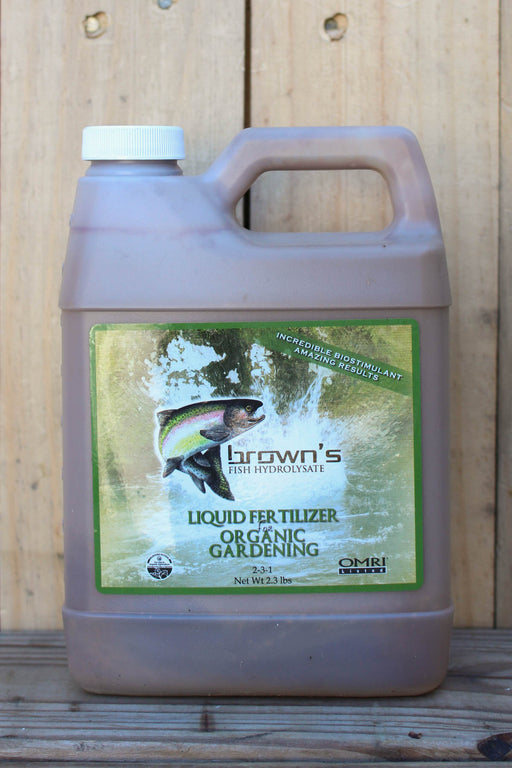 A large plastic jug of Brown's Liquid Fertilizer for Organic Gardening, filled with a brownish-purple liquid, set against a rustic wooden plank background.