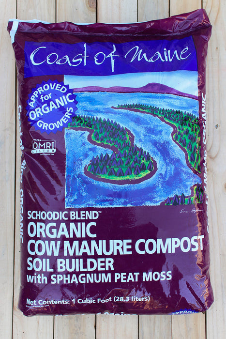 A bag of Coast of Maine Organic Cow Manure Compost Soil Builder, Schoodic Blend, shown on a light-colored wooden plank background. The packaging is dark purple with a central illustration of a winding blue river and green island.