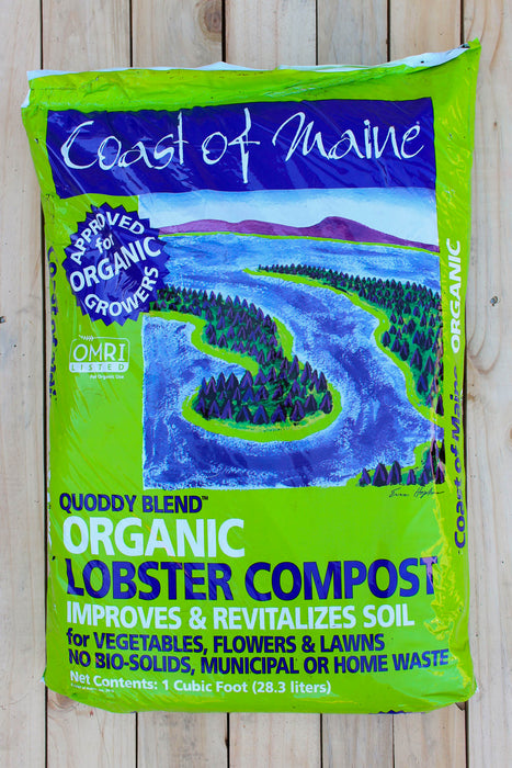 A bag of Coast of Maine Organic Lobster Compost Quoddy Blend, displayed flat on a light-colored wooden plank surface. The packaging is bright lime green with a dark blue landscape illustration of a winding river.