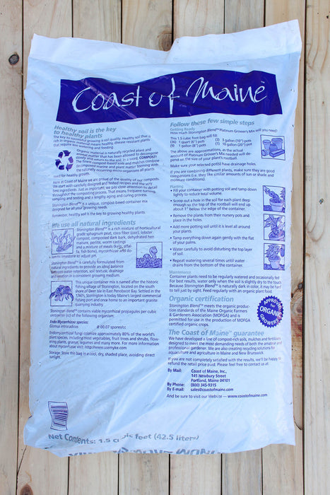 A white bag of Coast of Maine Polished Stone Flour, displayed flat on a light-colored wooden plank surface. The packaging features a prominent blue header with the brand name 'Coast of Maine' in white script, and extensive blue text and instructional diagrams detailing the product's use and ingredients.