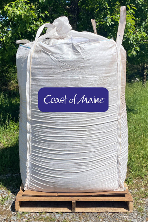 A large white bulk tote bag sitting on a wooden pallet outdoors, with green foliage and grass in the background. A purple label on the front reads 'Coast of Maine' in white script.