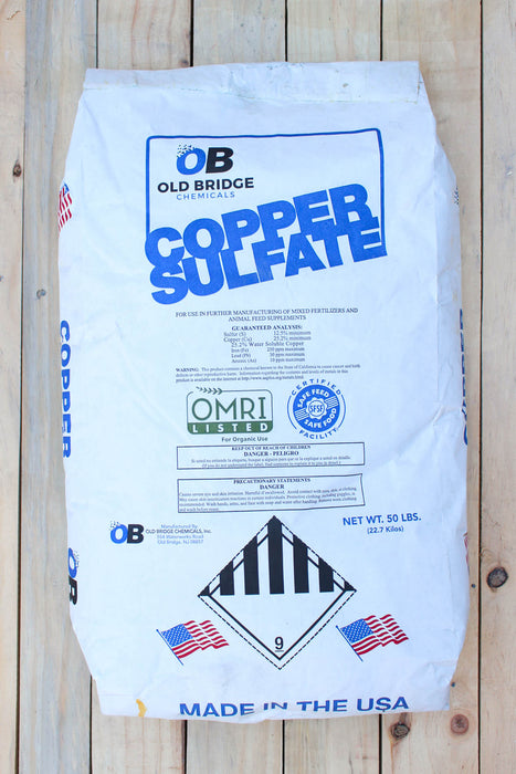 A 50 lb bag of Old Bridge Copper Sulfate fertilizer, featuring a white bag with blue and black text and graphics, placed on a light-colored wooden plank surface.