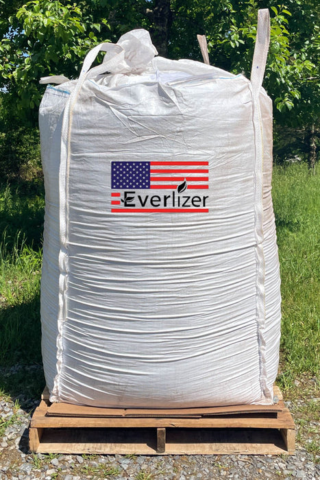 A large white bulk tote of Everlizer fertilizer sits on a wooden pallet outdoors, with green grass and trees in the background. The bag features a prominent red, white, and blue American flag logo with the brand name 'Everlizer' printed on the front.