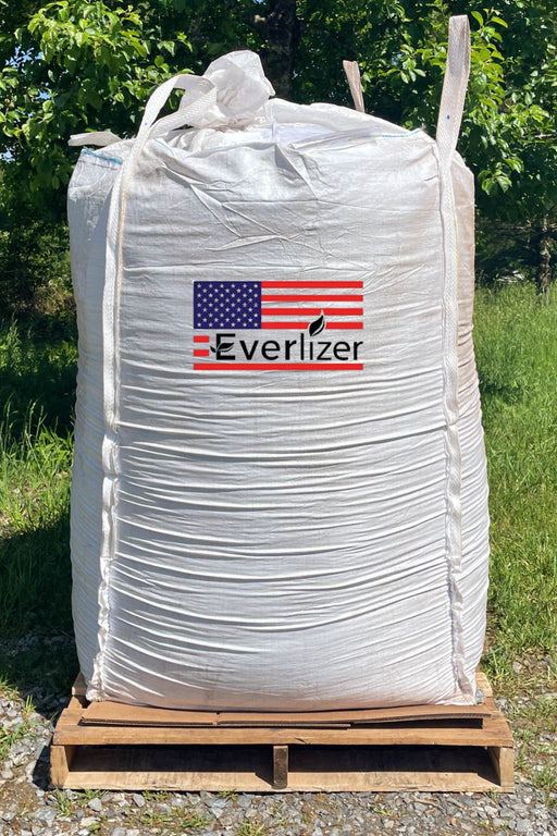 A large white bulk tote of Everlizer fertilizer sits on a wooden pallet outdoors, with green grass and trees in the background. The bag features a prominent red, white, and blue American flag logo with the brand name 'Everlizer' printed on the front.