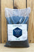 A large clear plastic bag filled with dark charcoal-colored Ferti Organic Soluble Seaweed Extract Plus, featuring a white and blue label, set against a rustic wooden plank background.