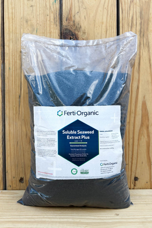 A large clear plastic bag filled with dark charcoal-colored Ferti Organic Soluble Seaweed Extract Plus, featuring a white and blue label, set against a rustic wooden plank background.