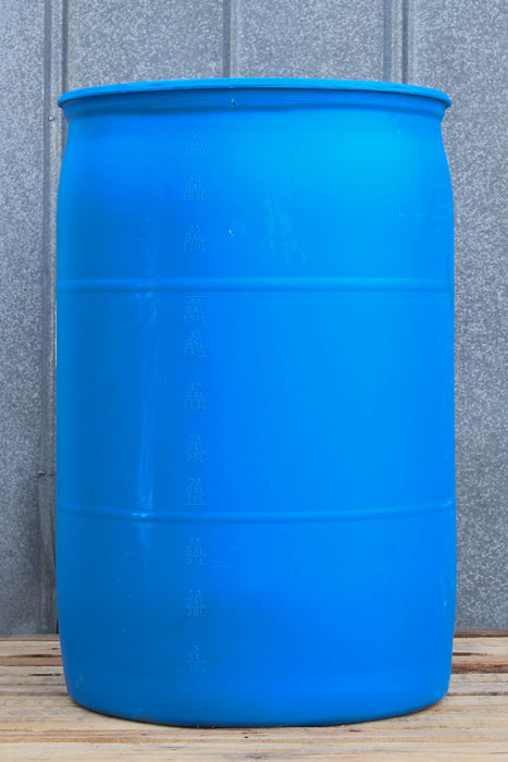 A large blue plastic barrel or drum with horizontal reinforcing rings, standing on a wooden surface against a gray corrugated metal wall.