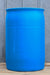 A large blue plastic barrel or drum with horizontal reinforcing rings, standing on a wooden surface against a gray corrugated metal wall.