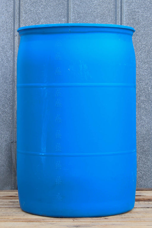 A large blue plastic barrel or drum with horizontal reinforcing rings, standing on a wooden surface against a gray corrugated metal wall.