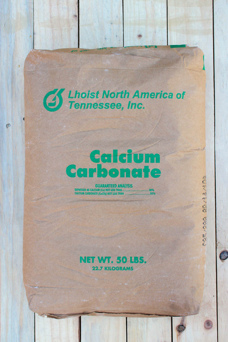 A 50 lbs brown paper bag of Calcium Carbonate, featuring green text and logo, placed flat on a light-colored wooden plank background.