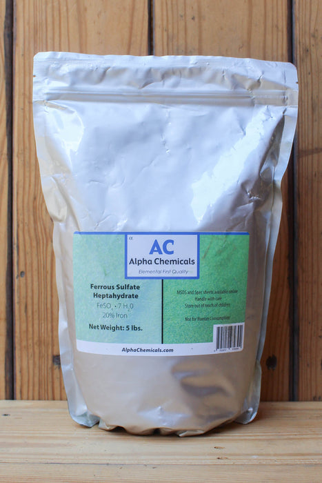 A 5 lb bag of Alpha Chemicals Ferrous Sulfate Heptahydrate, shown standing upright on a wooden surface against a wooden plank background.