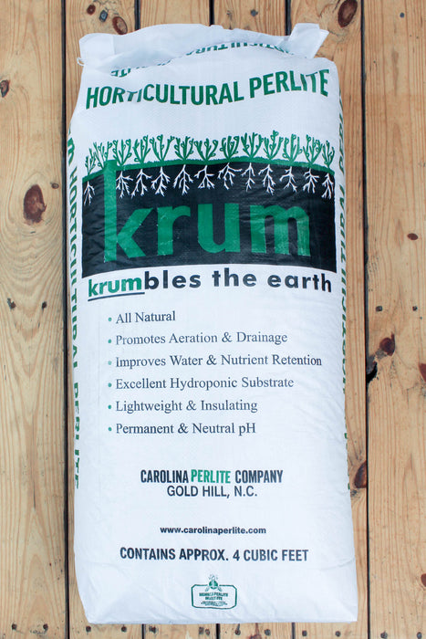 A large white bag of Krum Horticultural Perlite placed on a light brown wooden plank surface. The bag features green and black branding with text detailing its benefits as a natural, lightweight, and aeration-promoting substrate.