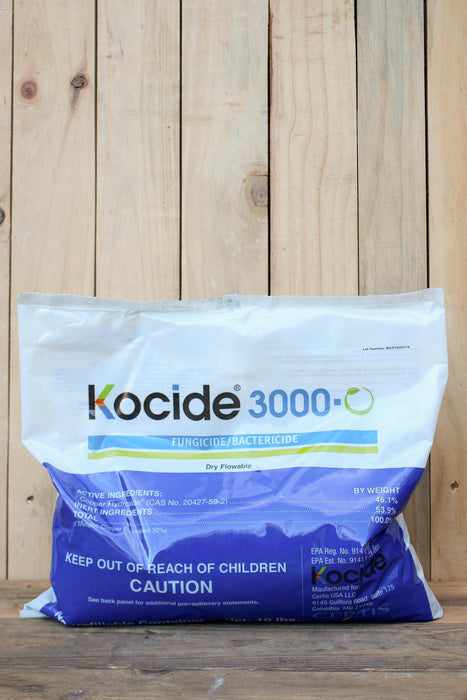 A large white and blue bag of Kocide 3000 fungicide and bactericide standing on a wooden surface against a light wood plank background.