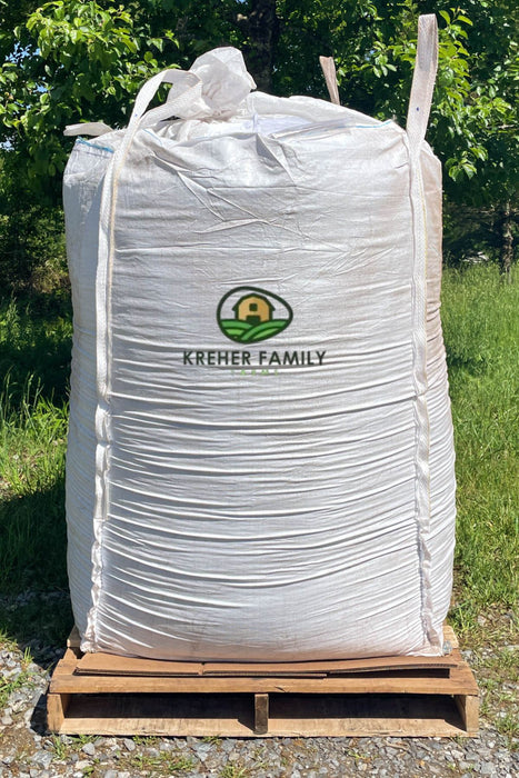 A large white bulk tote with the Kreher Family Farms logo, sitting on a wooden pallet outdoors on a grassy area with trees in the background.