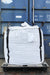 A large white tote bag with black webbing straps, resting on a black pallet in front of a blue shipping container.