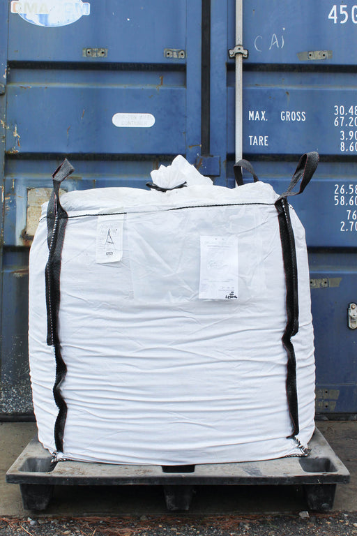A large white tote bag with black webbing straps, resting on a black pallet in front of a blue shipping container.