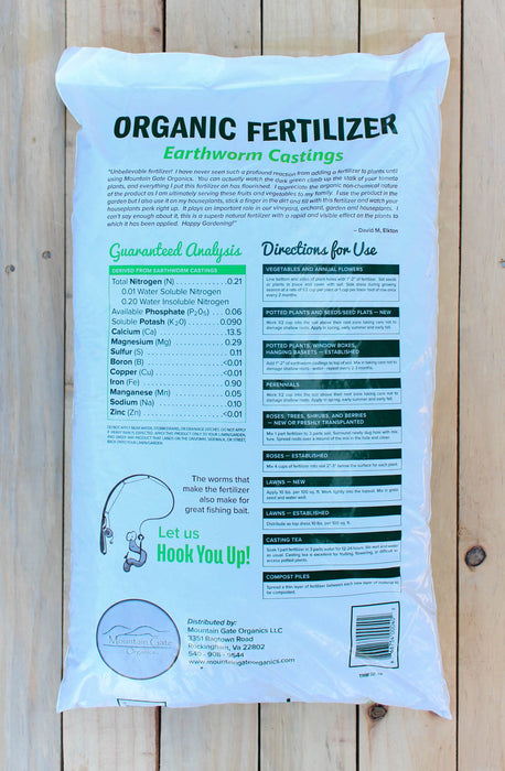 A large white bag of Earthworm Castings Organic Fertilizer lies flat on a light-colored wooden plank surface. The back of the bag features green and black text detailing the product's guaranteed analysis and directions for use, with a small illustration of a fishing line hook and a circular logo near the bottom.
