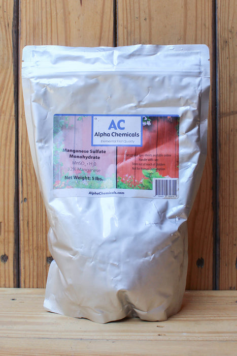 A large white stand-up pouch of Alpha Chemicals Manganese Sulfate Monohydrate standing on a wooden surface against a wooden plank background.