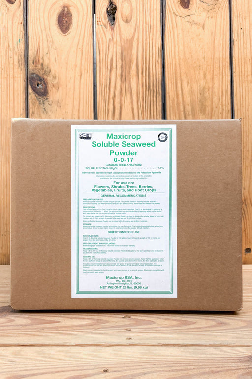 A brown cardboard box of Maxicrop Soluble Seaweed Powder with a white label, set against a rustic wooden plank background.