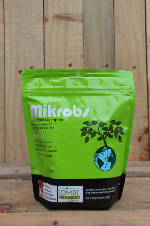A green stand-up pouch of Mikrob's Microbial Superpack with a black bottom section, photographed against a rustic wooden plank background.