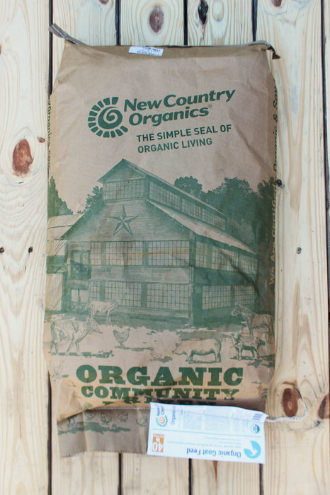 A brown paper bag of New Country Organics goat feed, featuring a green printed illustration of a barn and goats, laid flat on a light-colored wooden plank background.