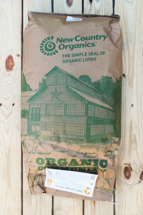 A large brown paper bag of New Country Organics Organic Livestock feed lies on a light wood plank surface. The bag features green printed branding, a barn illustration, and a white shipping label attached at the bottom.