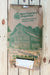A large brown paper bag of New Country Organics Organic Livestock feed lies on a light wood plank surface. The bag features green printed branding, a barn illustration, and a white shipping label attached at the bottom.