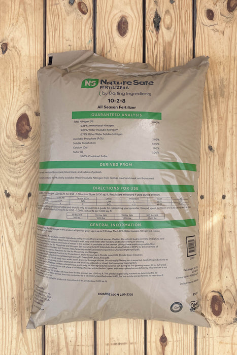 A large bag of Nature Safe fertilizer, 10-2-8 All-Season Formula, lying flat on a light-colored wooden plank surface.