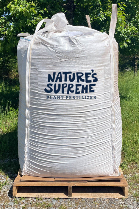 A large white plastic tote of Nature's Supreme Plant Fertilizer sitting on a wooden pallet outdoors on a gravel surface with green grass and trees in the background.