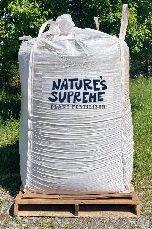 A large white plastic tote of Nature's Supreme Plant Fertilizer sitting on a wooden pallet outdoors on a gravel surface with green grass and trees in the background.