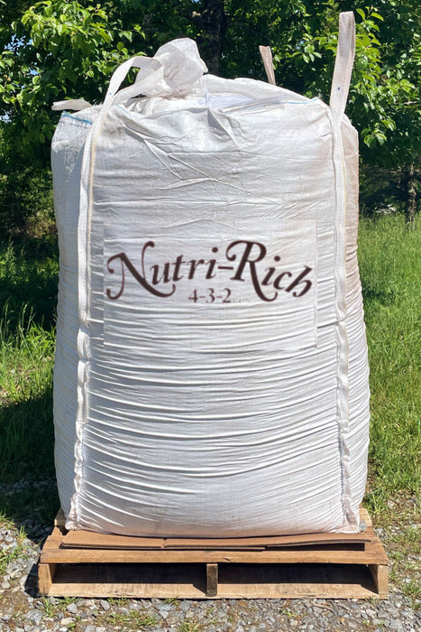A large white plastic bulk tote of Nutri-Rich 4-3-2 fertilizer sits on a wooden pallet outdoors on a gravel surface with green grass and trees in the background.