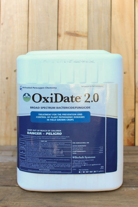 A large white plastic container of OxiDate 2.0 broad spectrum bactericide and fungicide, featuring a blue and white label, set against a vertical wood plank background.