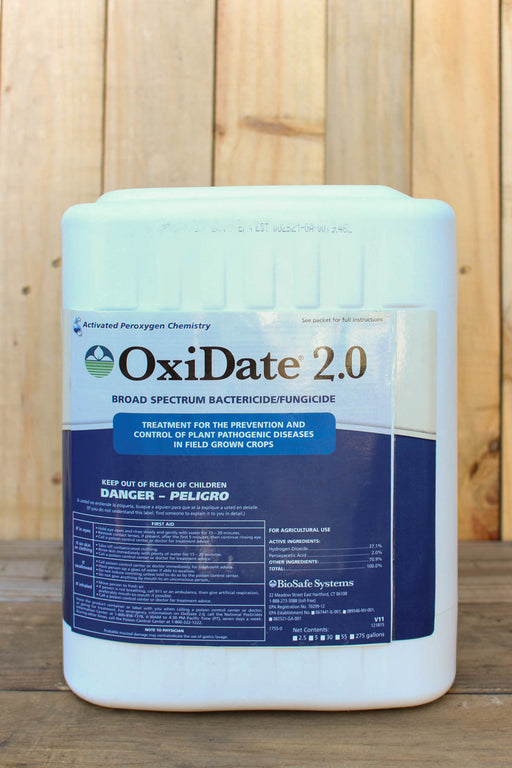 A large white plastic container of OxiDate 2.0 broad spectrum bactericide and fungicide, featuring a blue and white label, set against a vertical wood plank background.