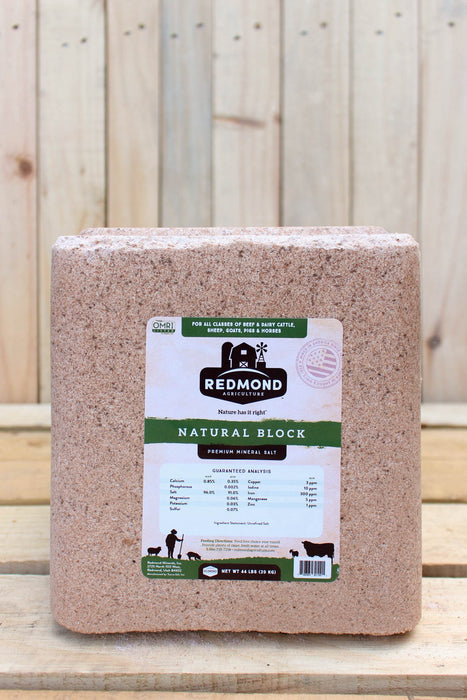 A large rectangular block of Redmond Natural Block premium mineral salt, featuring a white and green label, set against a rustic light wood plank background.