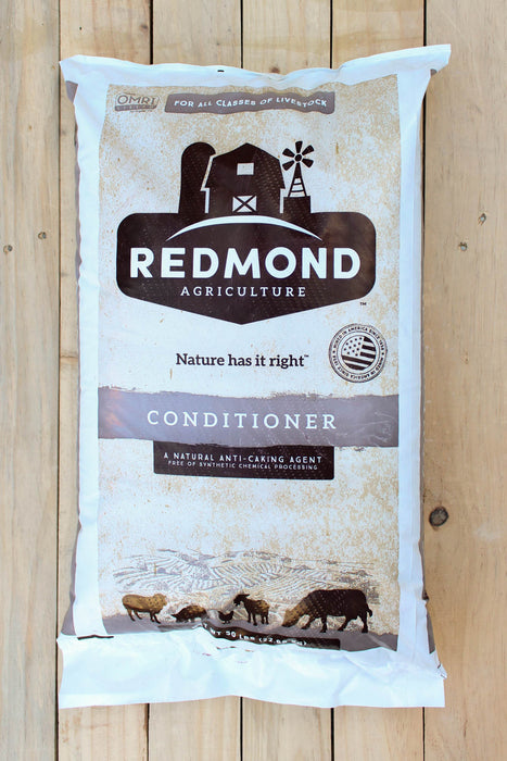 A large white and brown bag of Redmond Agriculture Conditioner, displayed flat on a light-colored wooden plank background.