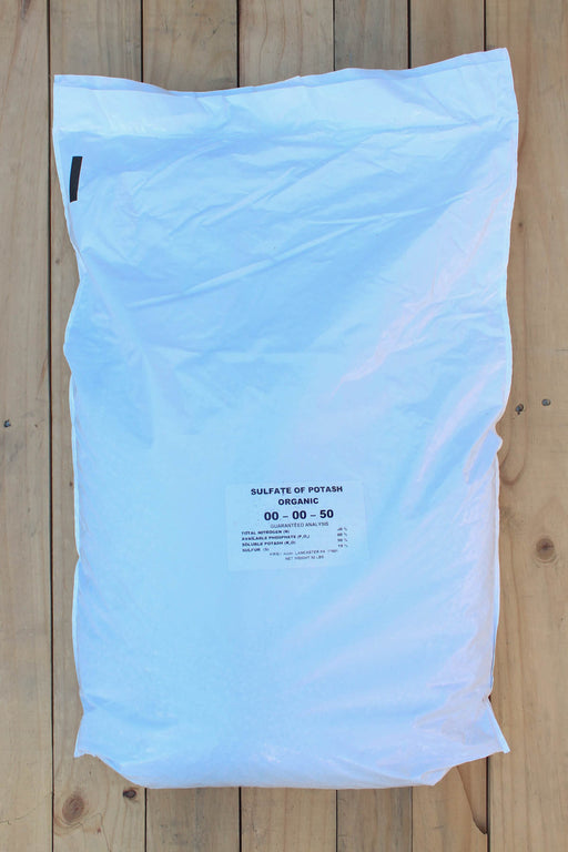 A large white plastic bag of Sulfate of Potash Organic fertilizer, labeled '00-00-50', laid flat on a light-colored wooden plank surface.