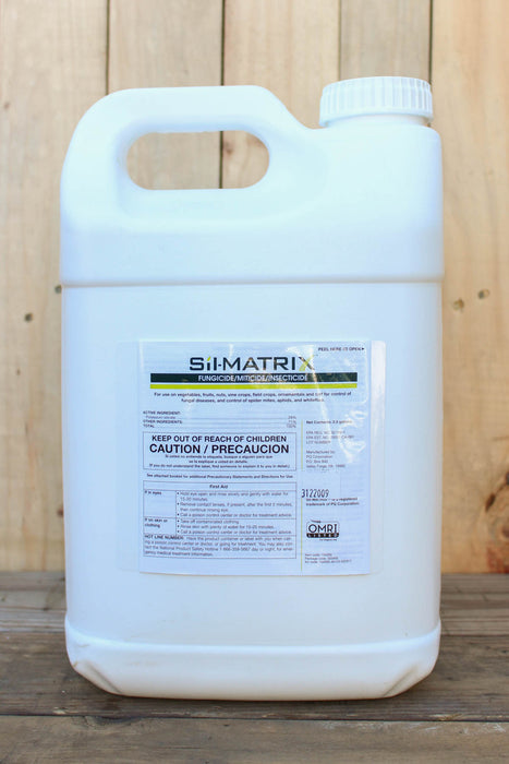 A large white plastic jug of Si-Matrix with a handle, standing on a wooden surface against a light wood plank background.