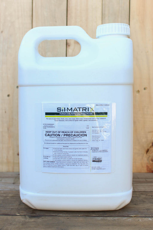 A large white plastic jug of Si-Matrix with a handle, standing on a wooden surface against a light wood plank background.