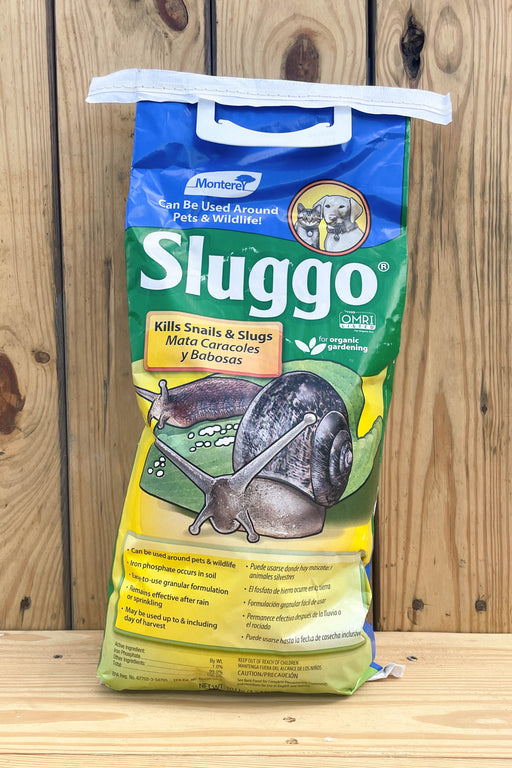 A bag of Sluggo slug and snail control treatment, featuring a blue and yellow design with illustrations of a snail and slug, standing on a wooden surface against a wooden plank background.