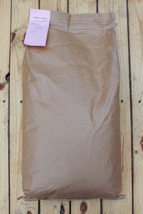 A large brown paper bag of Soybean Meal, with a pink paper label attached, laid flat on a light-colored wooden plank surface.