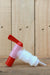 A plastic dispenser or faucet component with a red handle and red end, a translucent pink middle section, a white threaded body, and a clear tube, photographed on a wooden surface against a wood plank background.